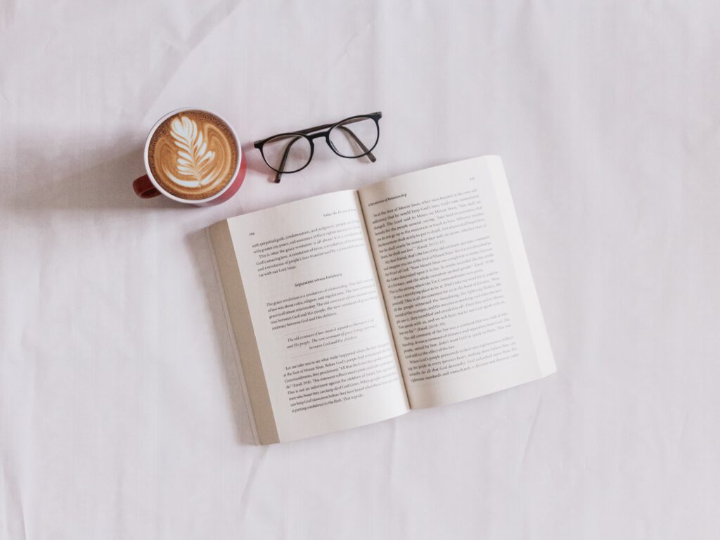 Open book, a cappuccino and a pair of reading glasses on white linen in the service page Italian editing services.